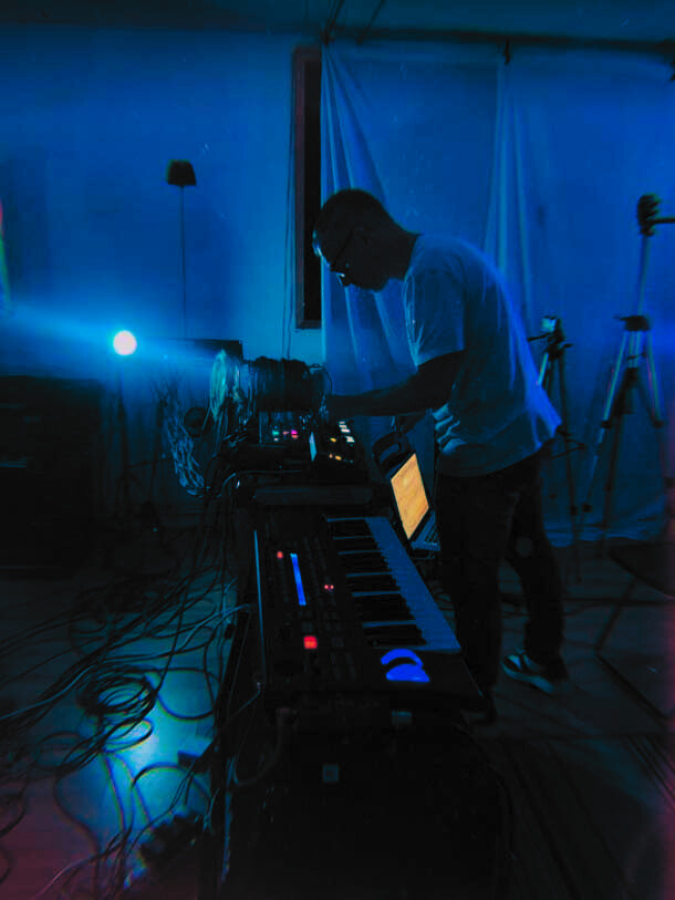 Dave hunched over a complicated set up of keyboards, synthesisers, drum machines and more. He's in the live room at VIBE Recording Studio, Manchester. The lighting is dark blue and all the equipment's lights glow brightly