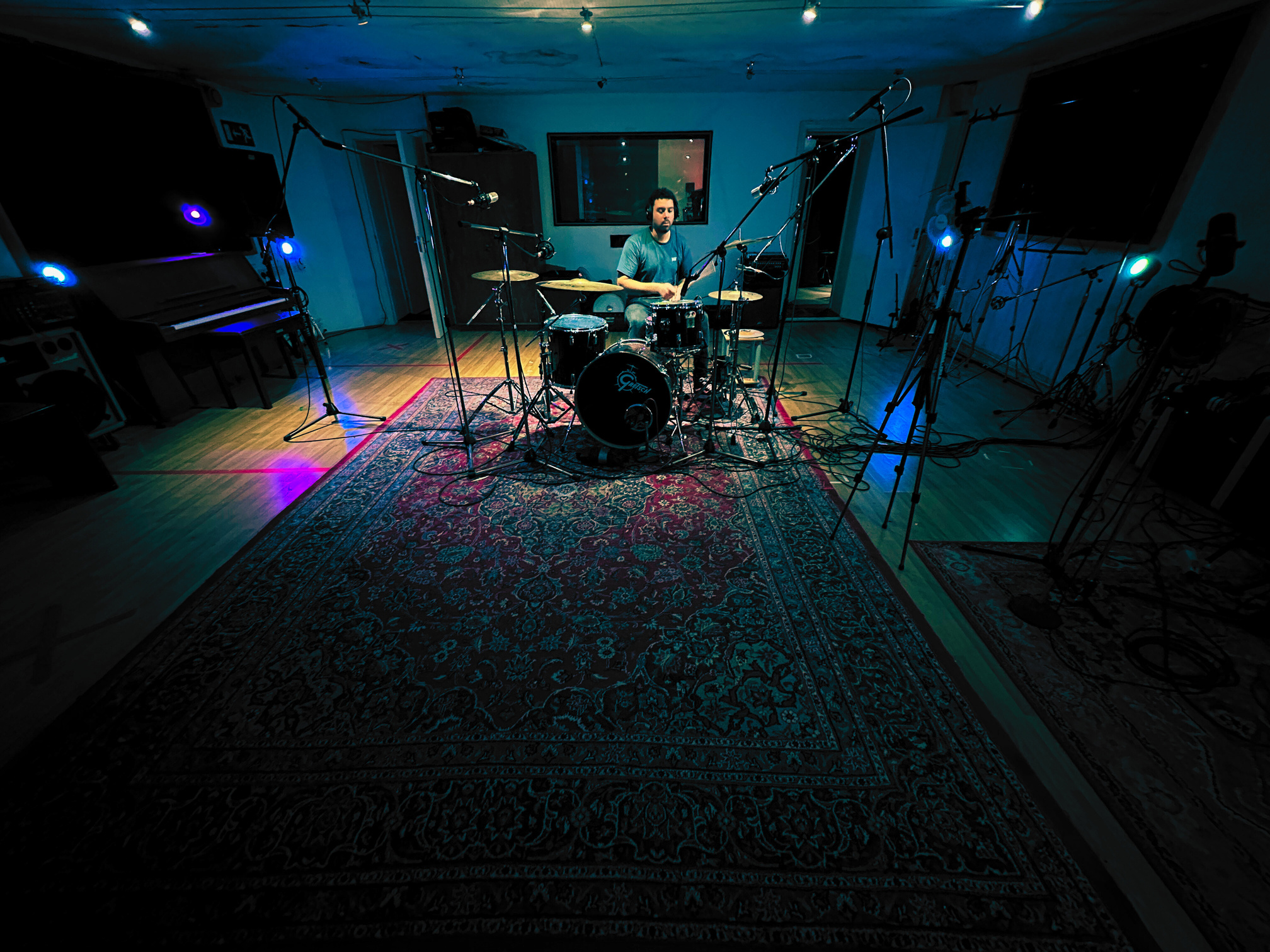 Lewis sits at the drum kit, sticks in hand playing a steady  beat in VIBE Recording Studio, Manchester. In the picture, the drumkit starts about two thirds of the way up, surrounded by hushed blue live room lighting and decorative long rug.