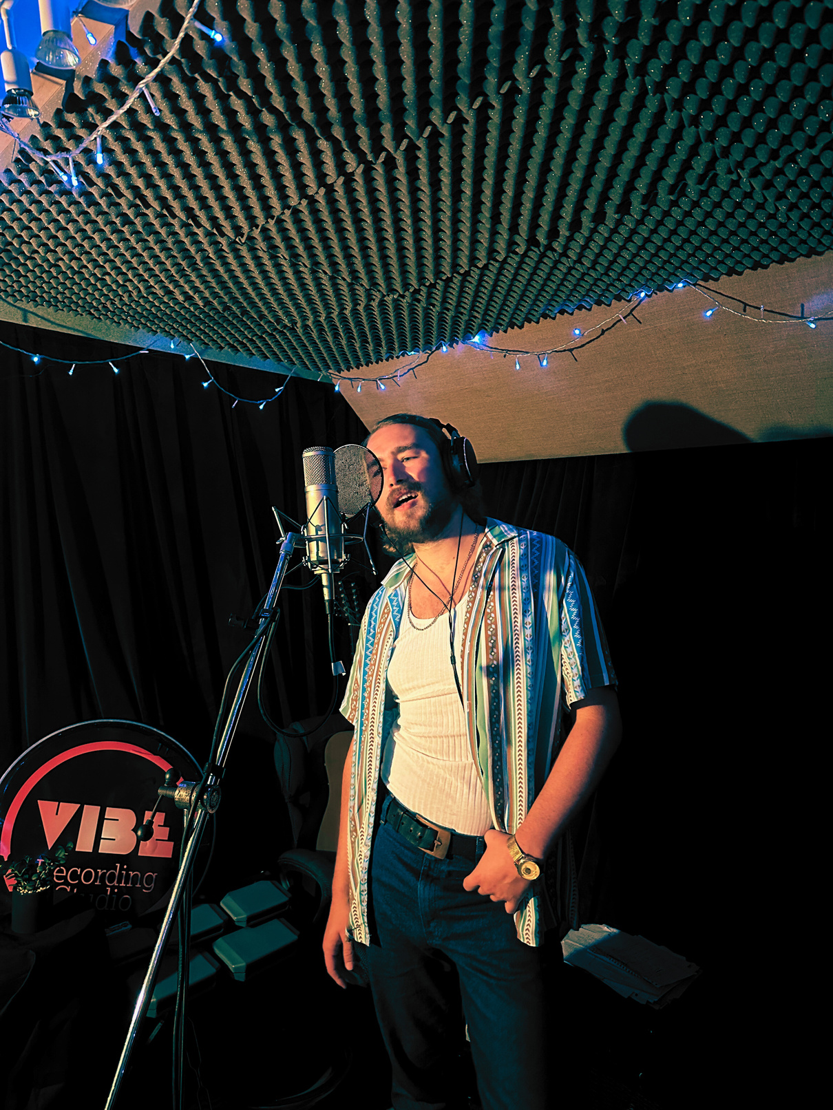 Ethan, stood one half to his side, one hand hanging off his belt, sings confidently into the VIBE Recording Studio microphone in Manchester. The vocal booth is laden with long curtains, acoustic treatment and fancy fairy lights round the top.